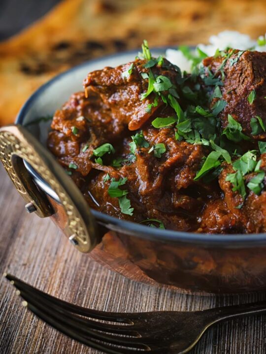 Portrait close up image of a lamb rogan josh curry served in a copper coated curry bowl with a naan bread
