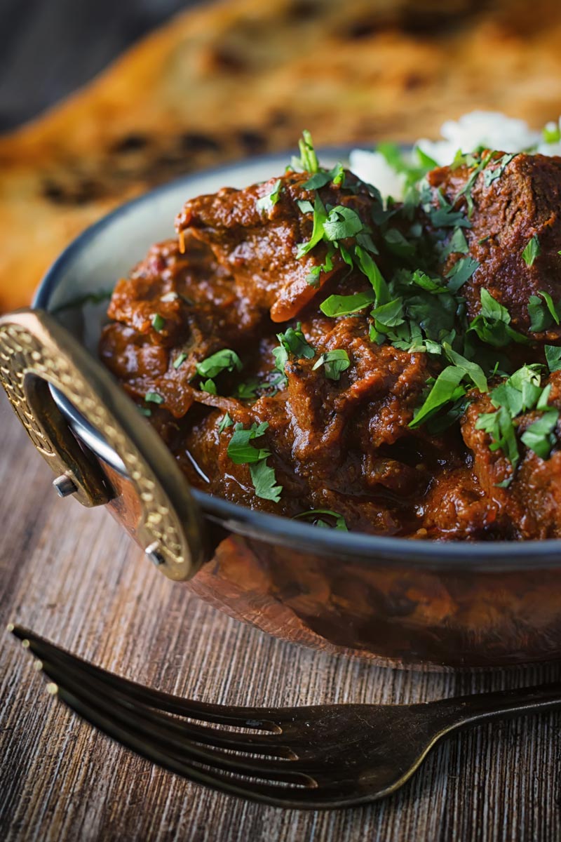 Portrait close up image of a lamb rogan josh curry served in a copper coated curry bowl with a naan bread
