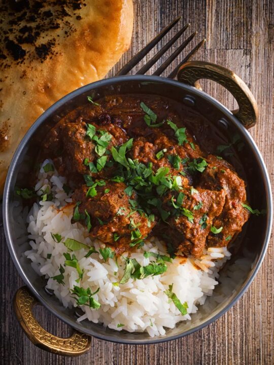 Portrait overhead image of a lamb rogan josh curry served in a copper coated curry bowl with a naan bread