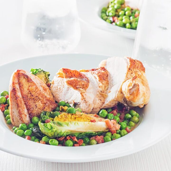 Square image of petits pois a la Francaise served in a white bowl with a sliced roast chicken breast on a bright backdrop with iced water