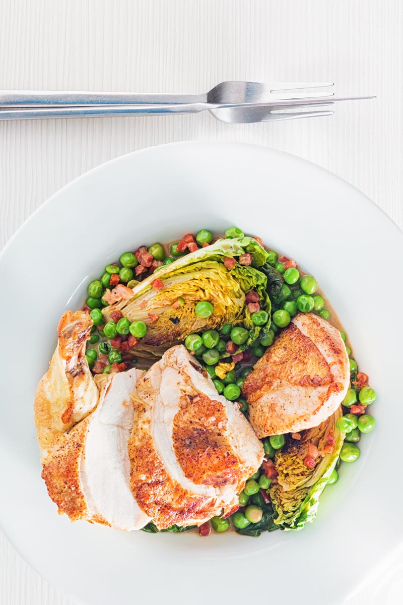 Portrait overhead image of petits pois a la Francaise served in a white bowl with a sliced roast chicken breast