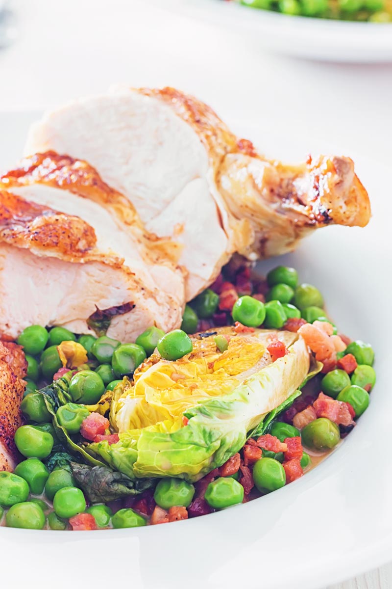Portrait close up image of petits pois a la Francaise served in a white bowl with a sliced roast chicken breast on a bright backdrop