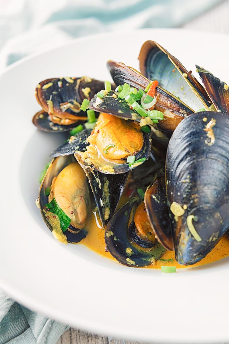 Portrait image of an Indian curry mussels served in a white bowl with a coconut broth and spring onions