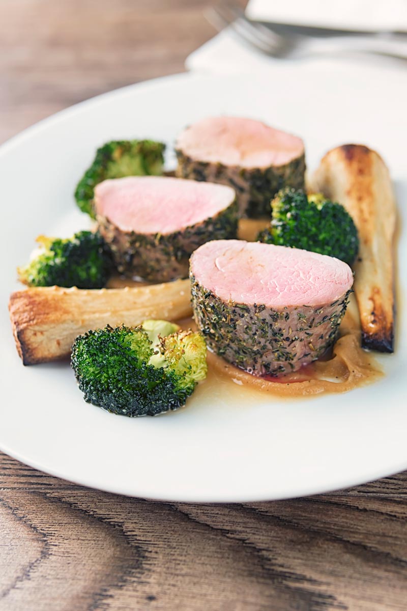 Portrait image of pink roast pork fillet served presented attractively on a white plate with apple puree, roast broccoli and parsnips and a cider sauce