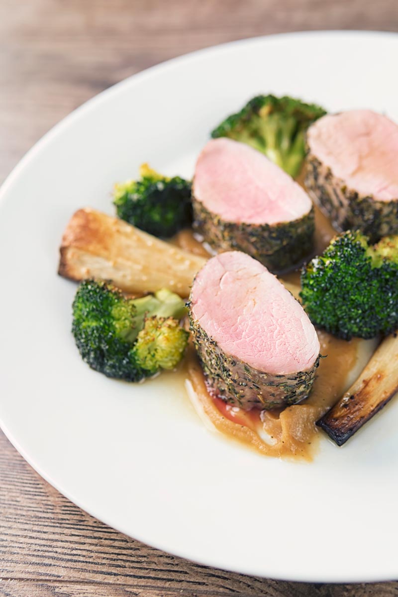 Portrait image of pink roast pork fillet served presented attractively on a white plate with apple puree, roast broccoli and parsnips and a cider sauce