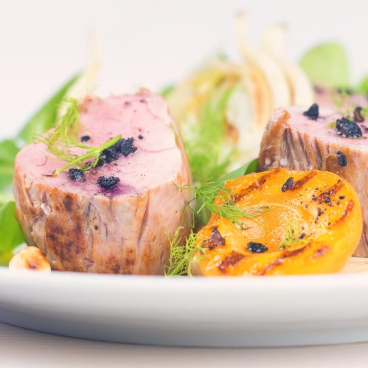 Square image of pink roast pork tenderloin with seared apricots and fennel served with Tasmanian pepper served ona white plate with purple linen