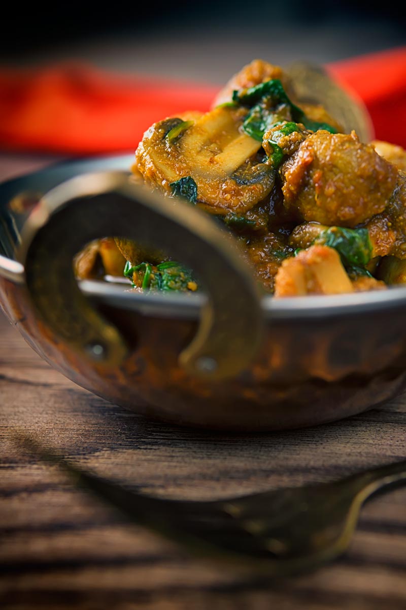Close up portrait image of a spinach and mushroom curry served in a copper bowl on a rustic wooden table with red linen and a vintage fork