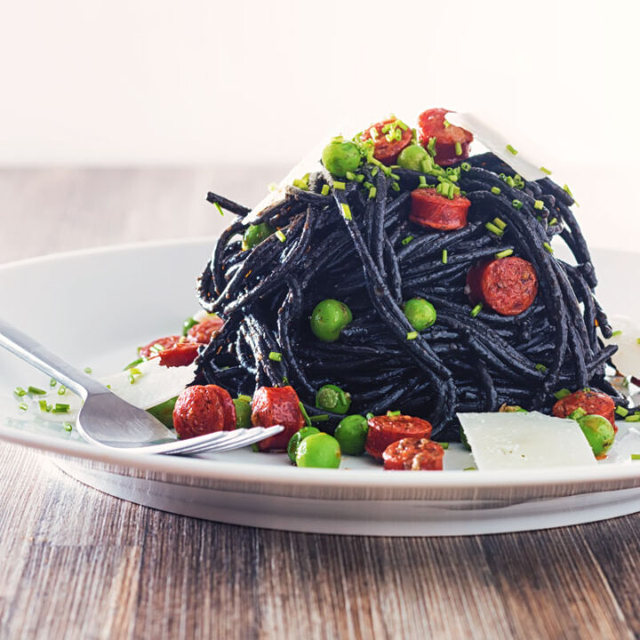 Square image of a tall pine of squid ink spaghetti on a white plate served with peas, pepperoni and parmesan cheese shavings