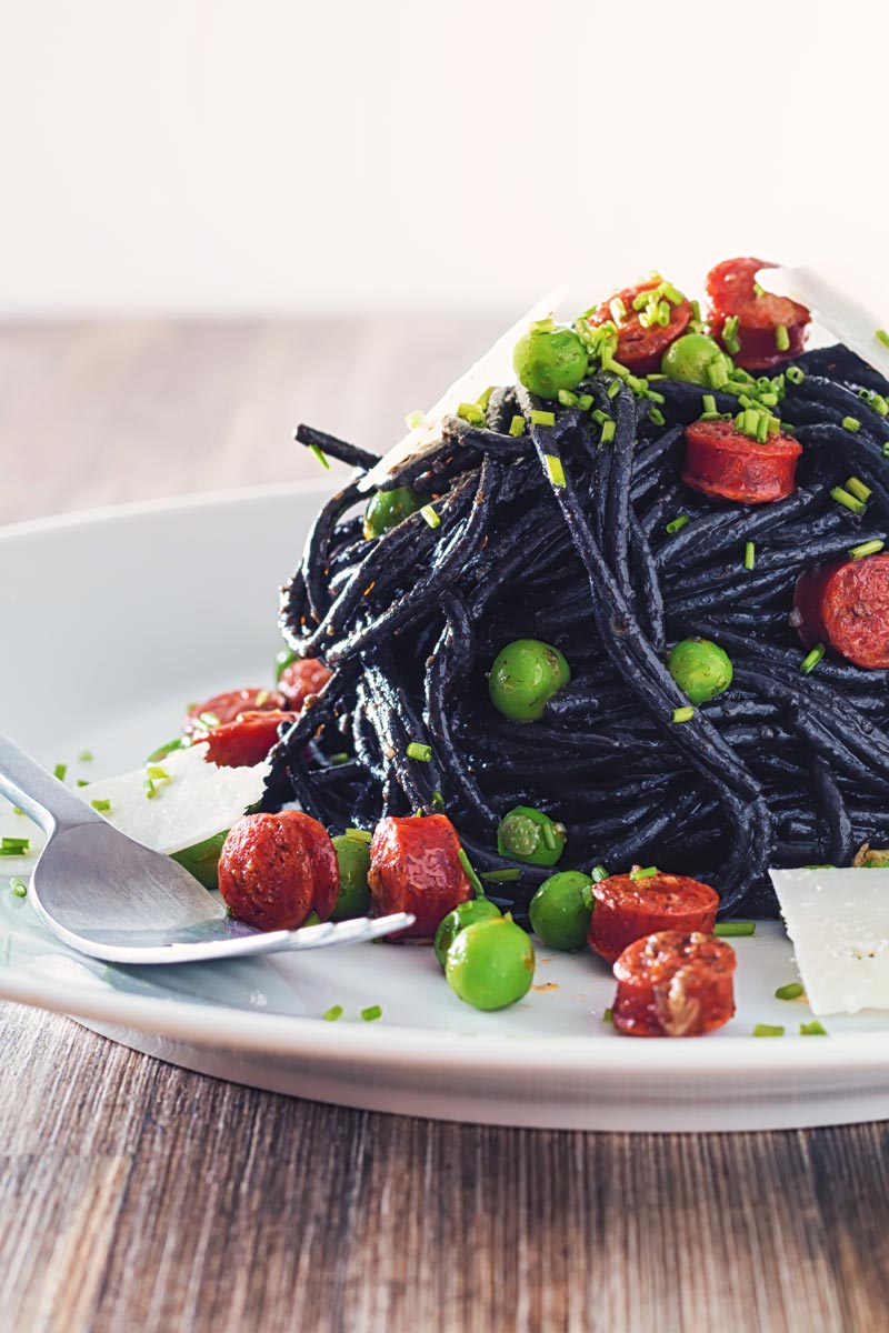 Portrait image of a tall pine of squid ink spaghetti on a white plate served with peas, pepperoni and parmesan cheese shavings