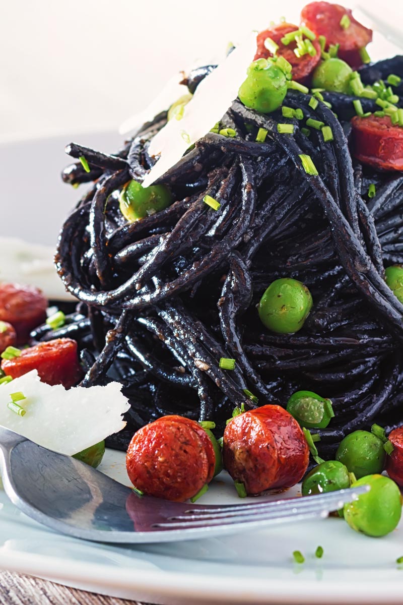 Close up portrait image of a tall pine of squid ink spaghetti on a white plate served with peas, pepperoni and parmesan cheese shavings