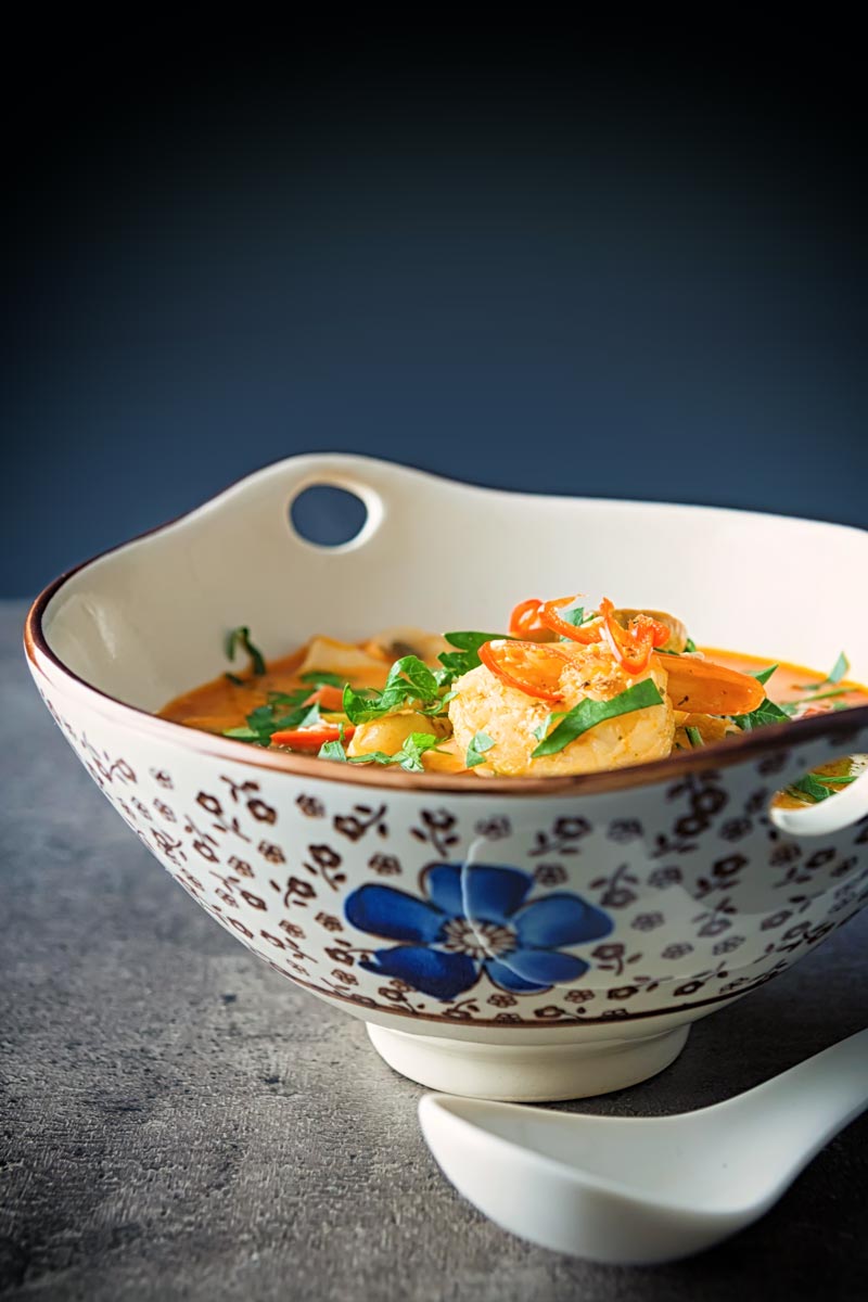 Portrait image of a Tom Yum Soup served in an Asian style soup bowl decorated with a blue flower in a dark setting