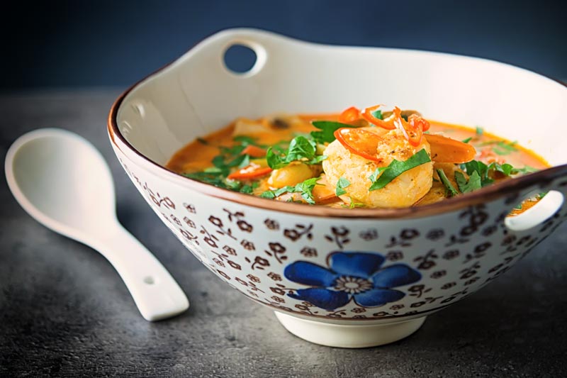 Landscape image of a Tom Yum Soup served in an Asian style soup bowl decorated with a blue flower in a dark setting