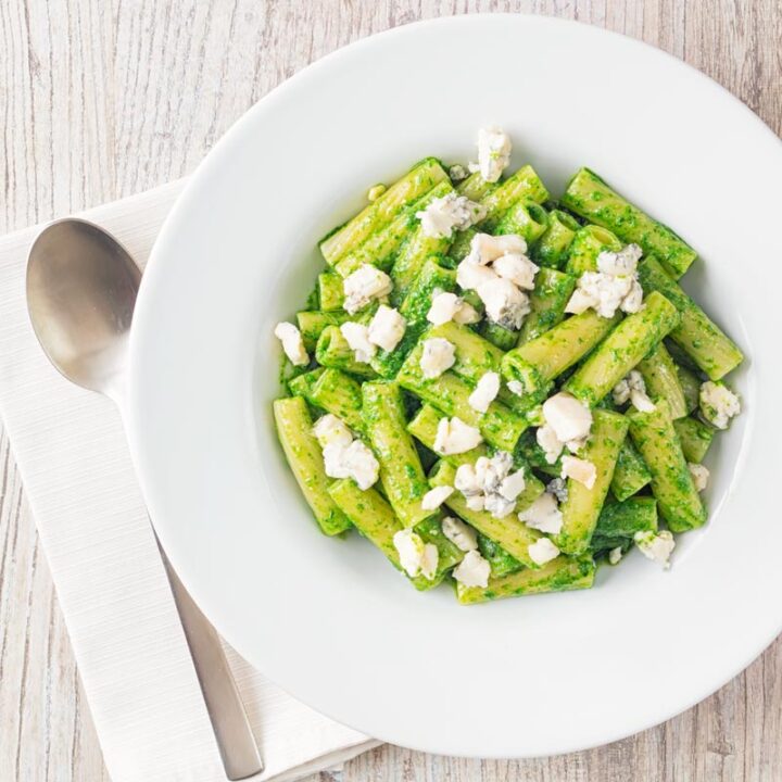 Square overhead image of wild garlic pesto pasta using rigatoni pasta garnished with gorgonzola cheese served in a wide white bowl