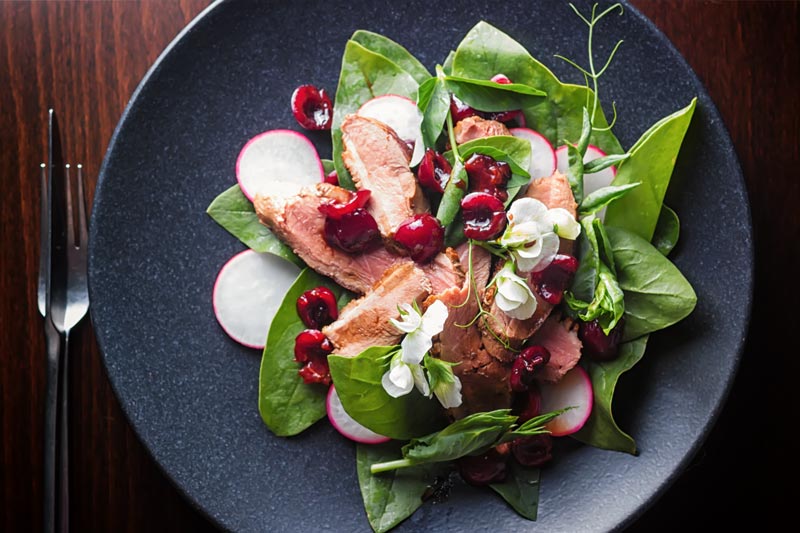 Landscape overhead image of a duck salad featuring sliced pan fried duck breast, Balsamic cherries, Spinach and Radish served on a black plate