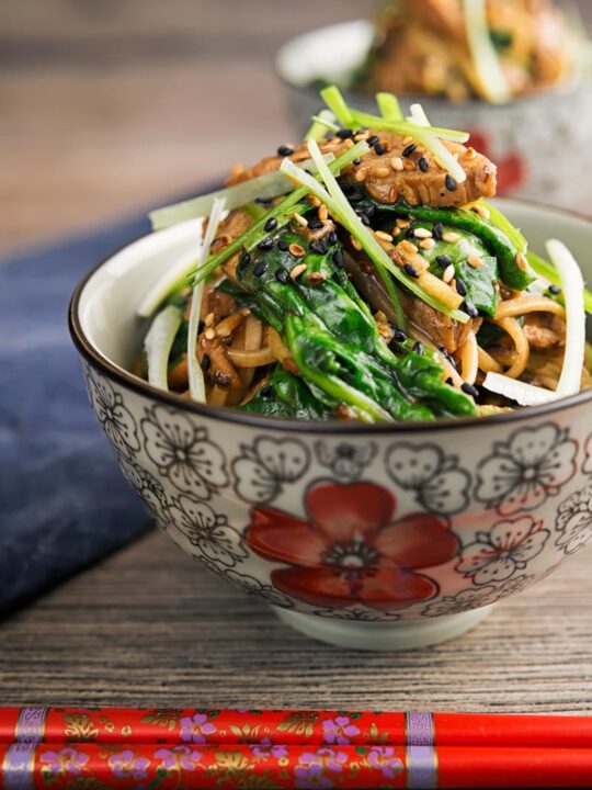 Square image of a pork stir fry with spinach and noodles served an Asian style bowl decorated with a red flower