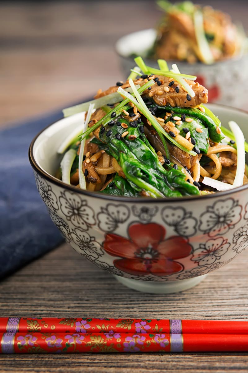 Square image of a pork stir fry with spinach and noodles served an Asian style bowl decorated with a red flower