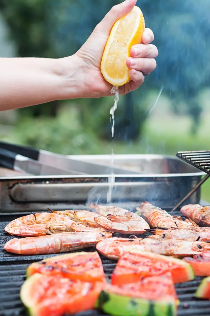 Portrait image of fresh lemon being squeezed over BBQ prawns or shrimp on a BBQ with watermelon