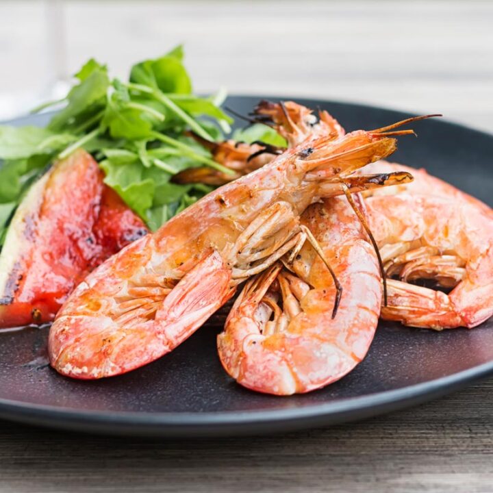 Square image of large BBQ prawns or shrimp with grilled watermelon served on a black plate