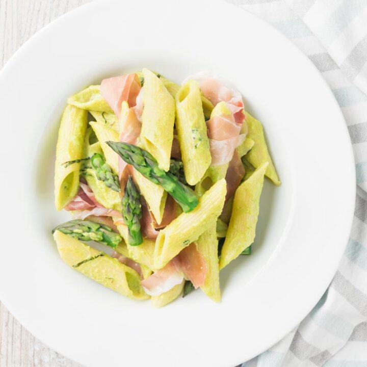 Square overhead image of penne asparagus pasta with asparagus spears and Parma ham in a white bowl with vintage cutlery