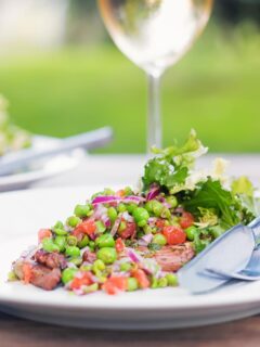Portrait image of a minted pea salsa served with lamb and a side salad on a white plate in a garden setting with a glass of wine.
