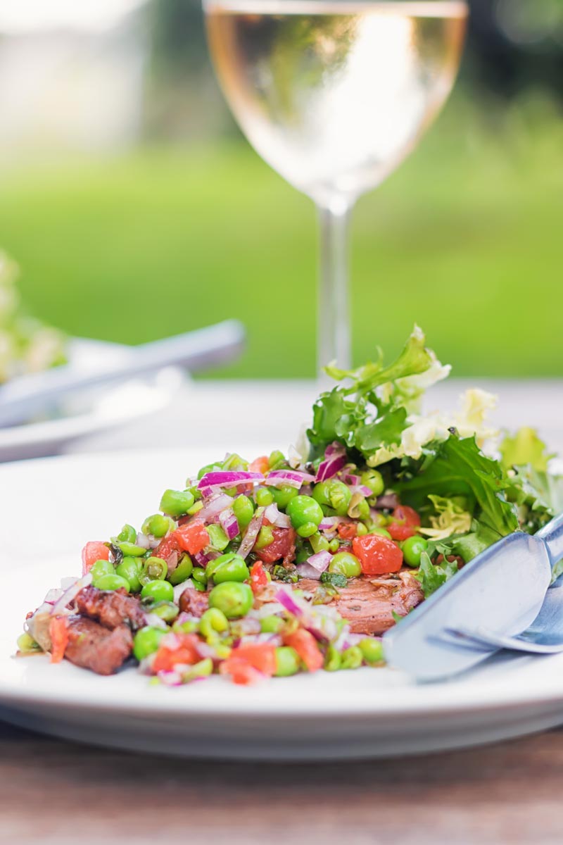 Portrait image of a minted pea salsa served with lamb and a side salad on a white plate in a garden setting with a glass of wine.