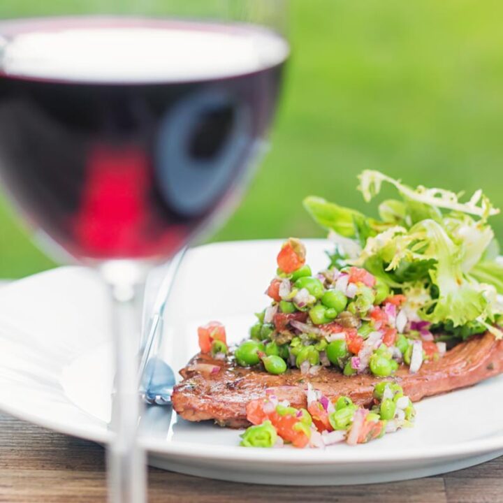 Square image of a minted pea salsa served with lamb and a side salad on a white plate in a garden setting with a glass of wine.