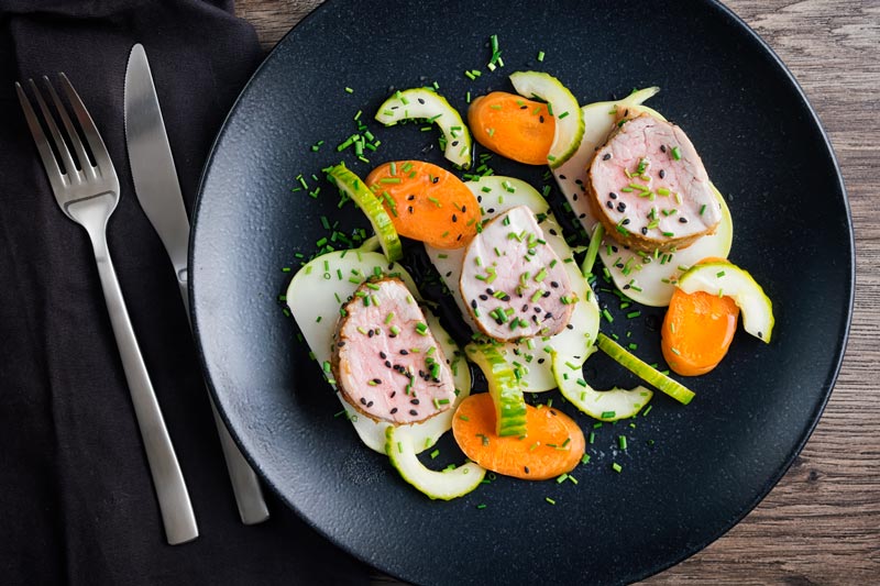 Landscape image of Miso and honey glazed pork tenderloin served on a black plate with pickled kohlrabi, carrots and cucumber