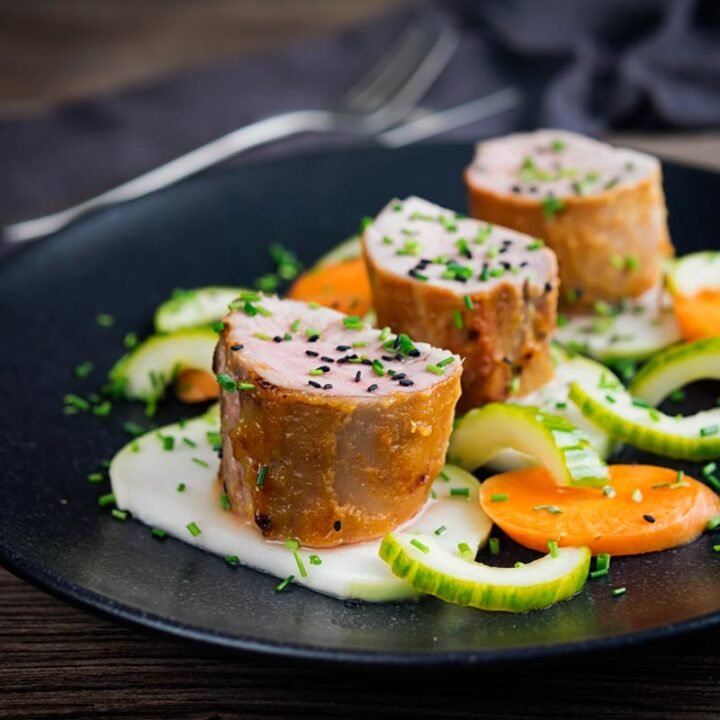Square image of Miso and honey glazed pork tenderloin served on a black plate with pickled kohlrabi, carrots and cucumber