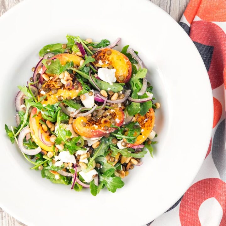 Square image of a seared peach and feta cheese salad with rocket, puy lentils and toasted pine nuts served in a white bowl