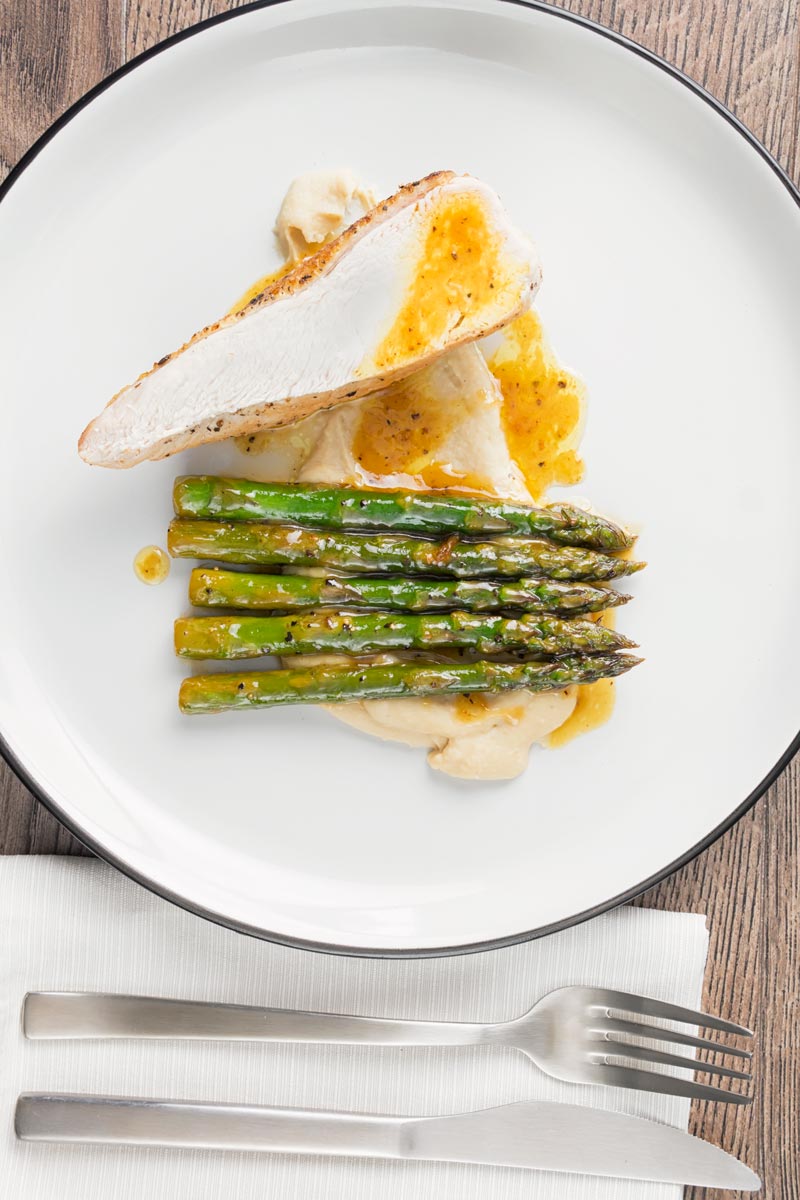 Portrait overhead image of a roast chicken breast served with white bean puree and orange asparagus served on a white plate with a black rim