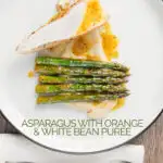Portrait overhead image of a roast chicken breast served with white bean puree and orange asparagus served on a white plate with a black rim with text