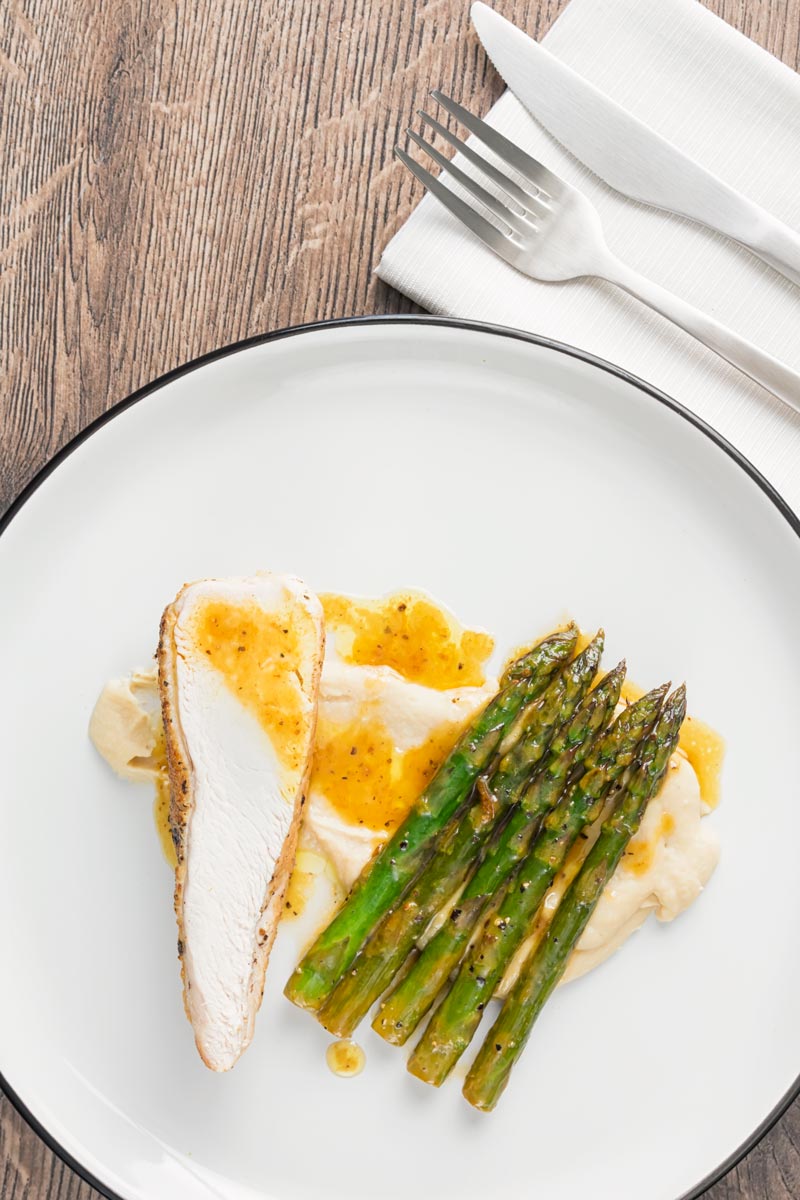 Portrait overhead image of a roast chicken breast served with white bean puree and orange asparagus served on a white plate with a black rim