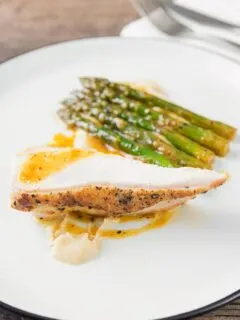 Portrait image of a roast chicken breast served with white bean puree and orange asparagus served on a white plate with a black rim