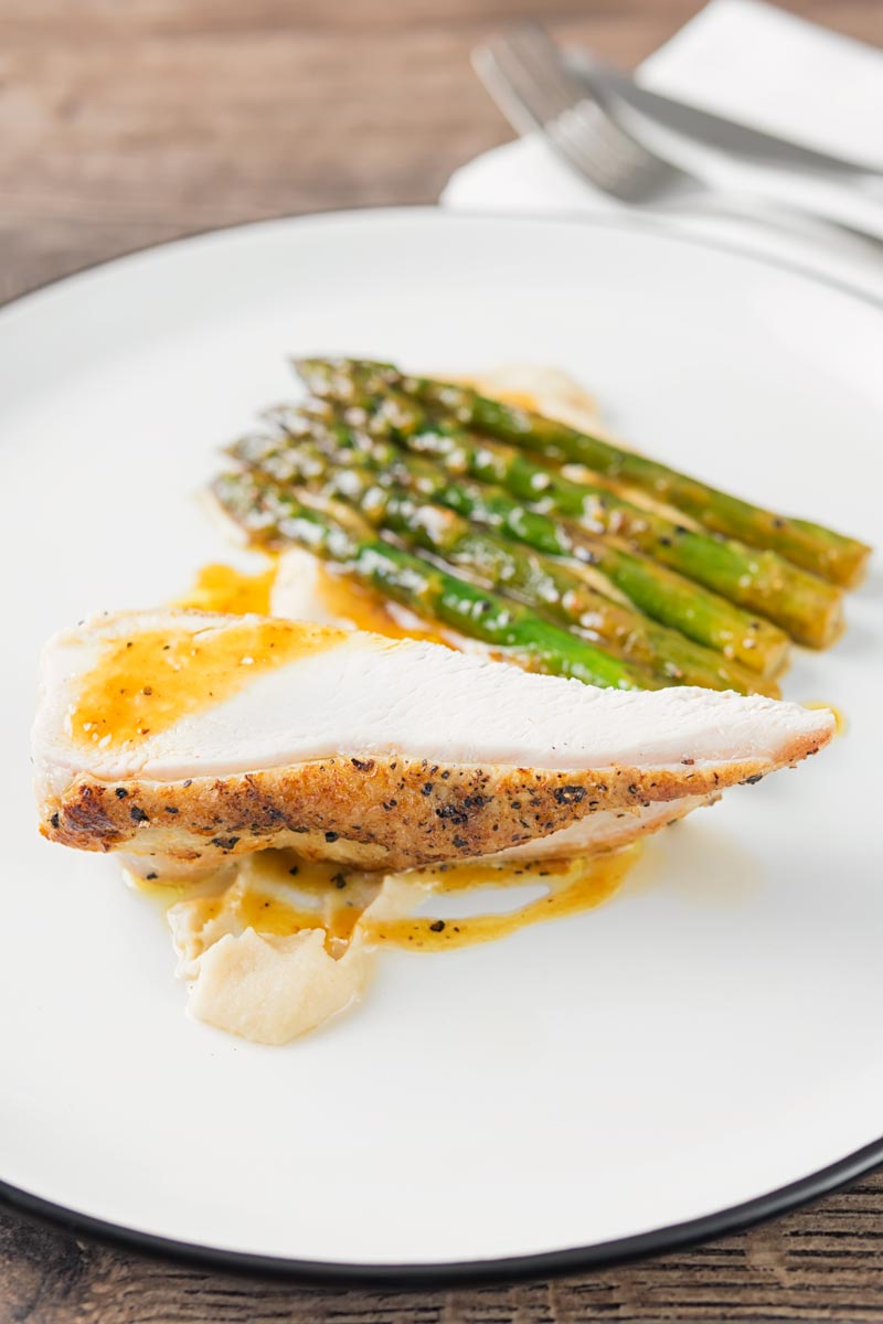 Portrait image of a roast chicken breast served with white bean puree and orange asparagus served on a white plate with a black rim