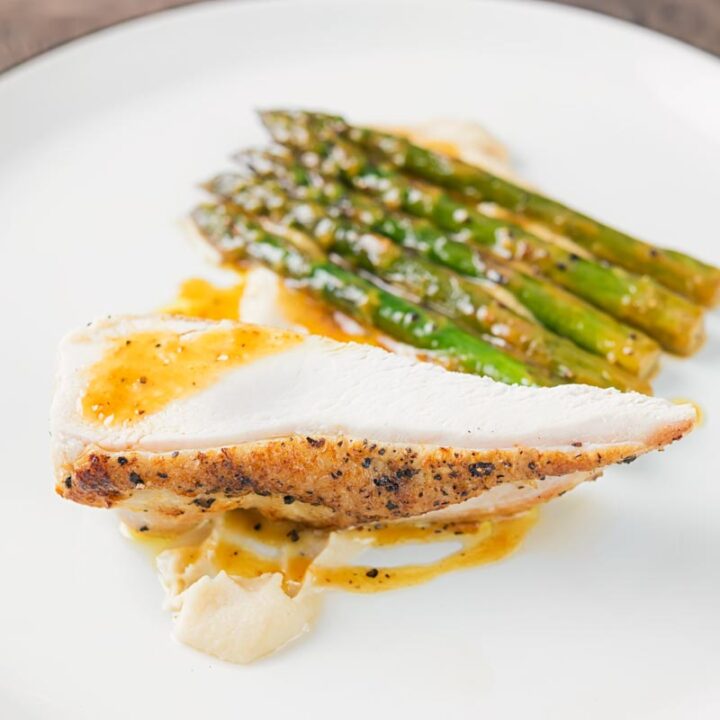 Sqaure image of a roast chicken breast served with white bean puree and orange asparagus served on a white plate with a black rim