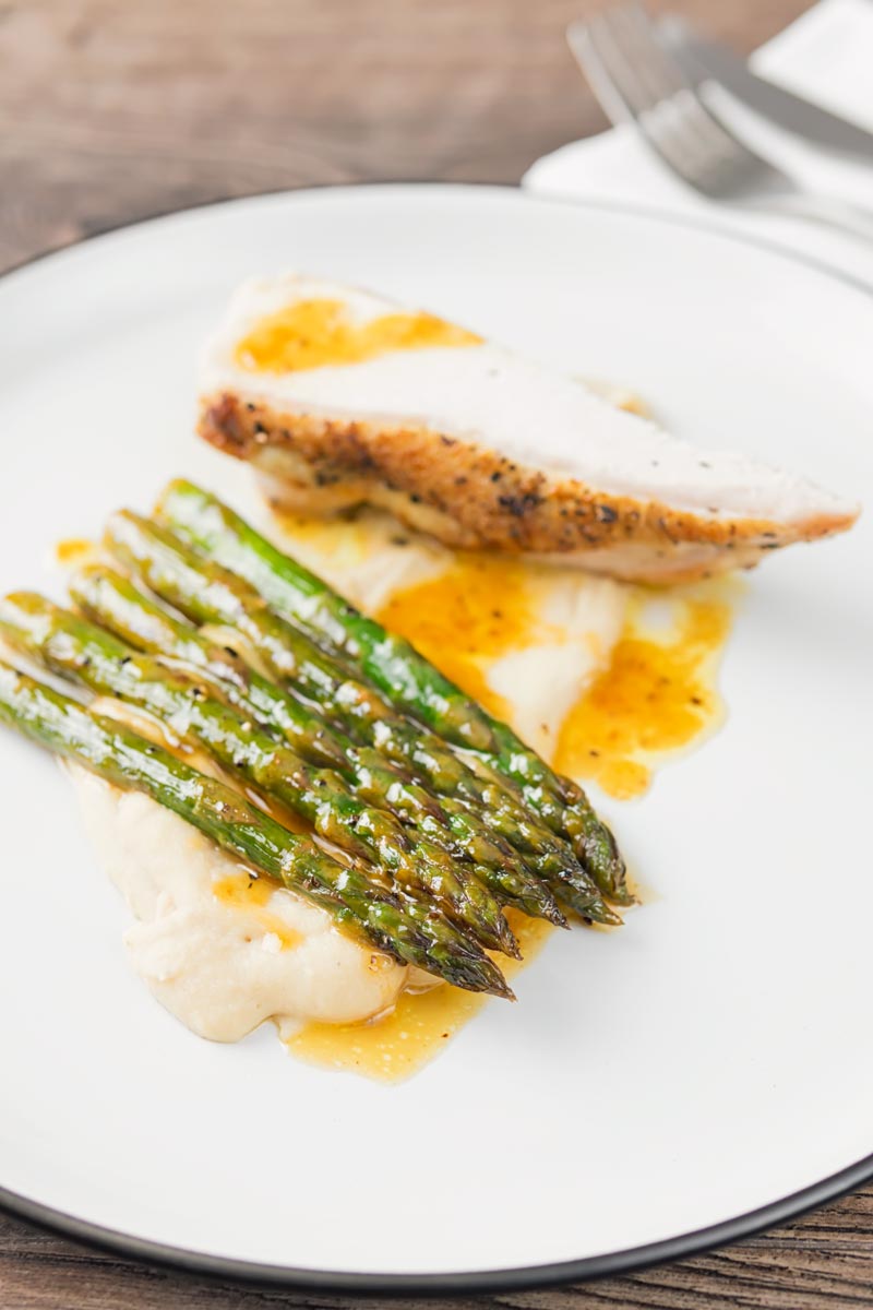 Portrait image of orange asparagus served with white bean puree and a roast chicken breast served on a white plate with a black rim