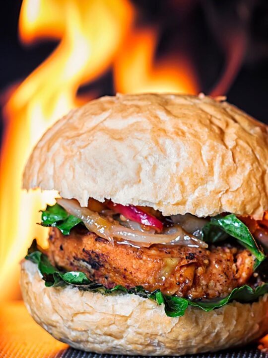 Portrait image of a spicy bean burger topped with an onion and peach chutney served on a bun with a flaming background