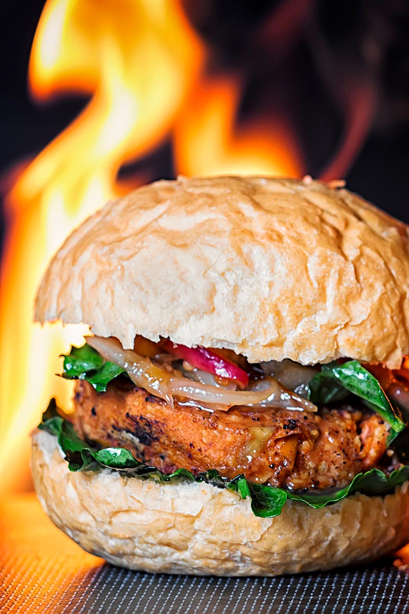Portrait image of a spicy bean burger topped with an onion and peach chutney served on a bun with a flaming background