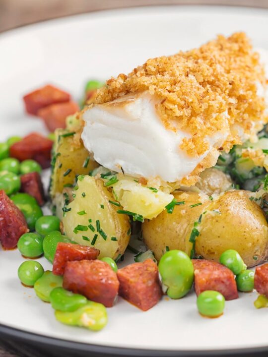 Portrait image of a baked cod fillet served on new potatoes served with peas, broad beans and chorizo sausage on a white plate
