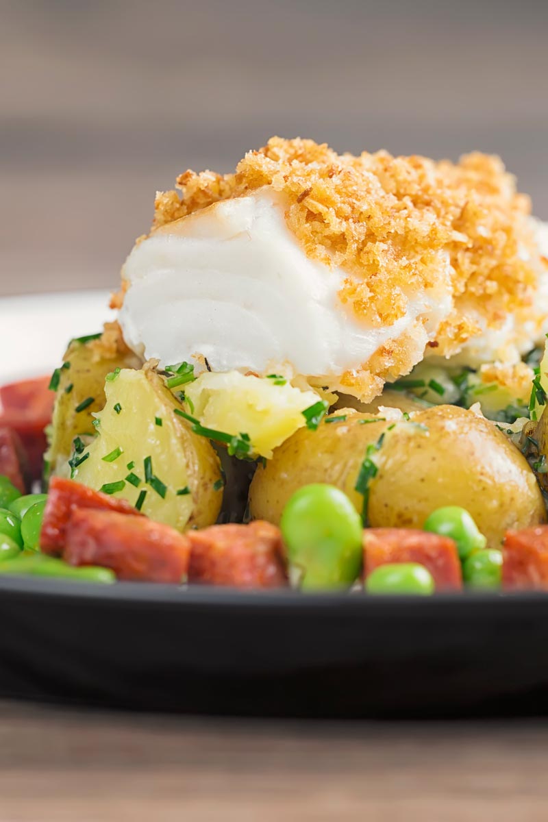 Portrait close up image of a baked cod fillet served on new potatoes served with peas, broad beans and chorizo sausage on a white plate
