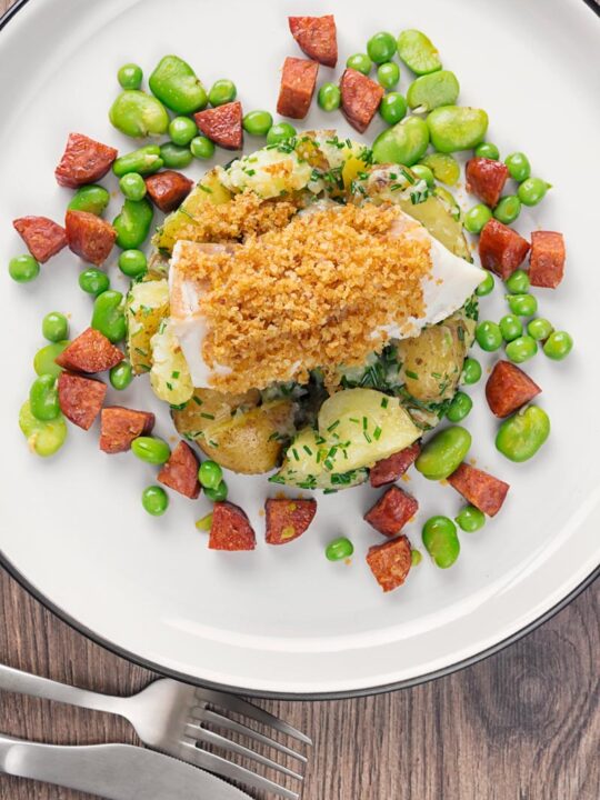 Portrait overhead image of a baked cod fillet served on new potatoes served with peas, broad beans and chorizo sausage on a white plate
