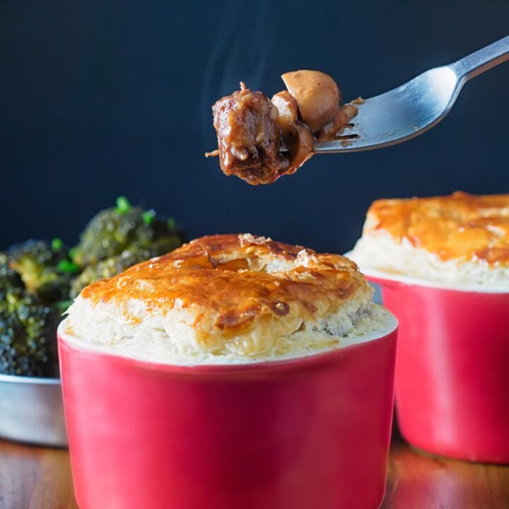 Square image of a beef and ale pie served pot pie style in a red pot with a fork removing steaming pie filling with text overlay