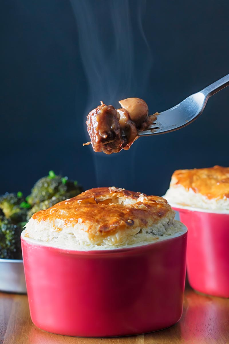 Portrait image of a beef and ale pie served pot pie style in a red pot with a fork removing steaming pie filling