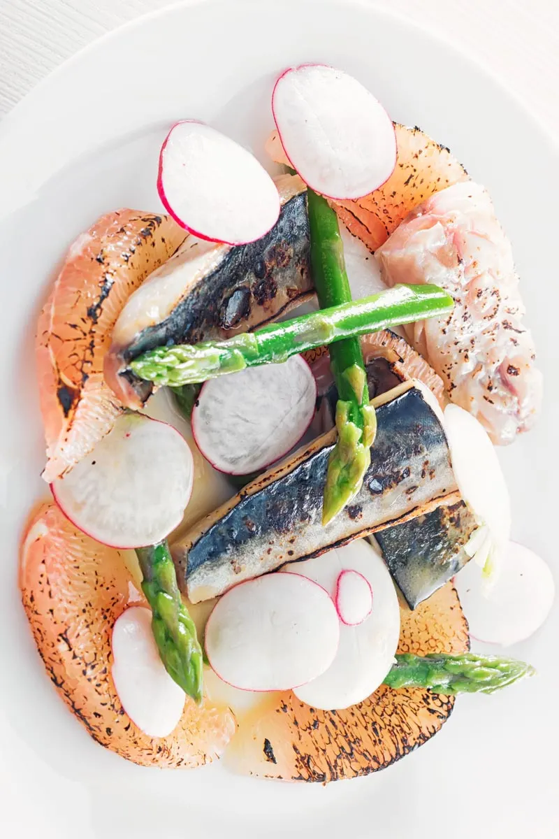 Portrait overhead image of cured mackerel served in a salad featuring asparagus spears and pomelo