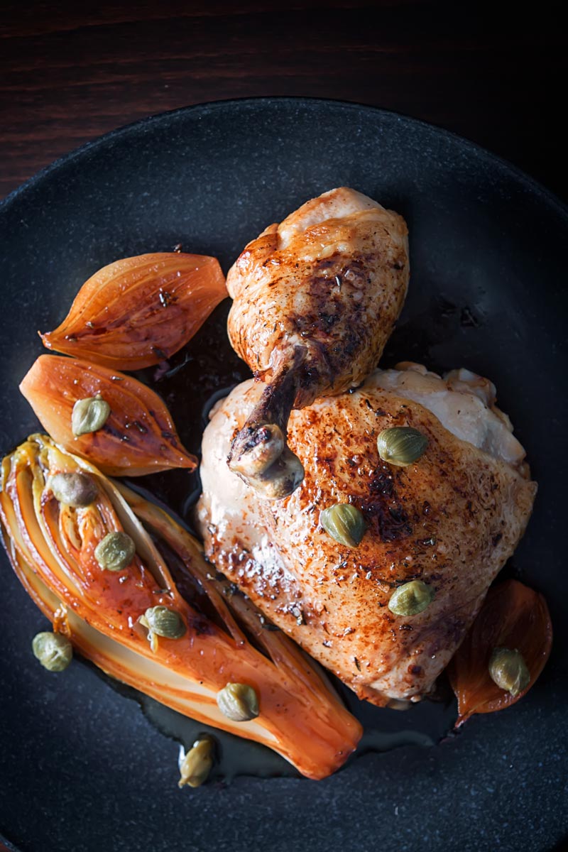Portrait overhead image of braised chicken legs served with Belgian Endive, shallots and capers on a black plate