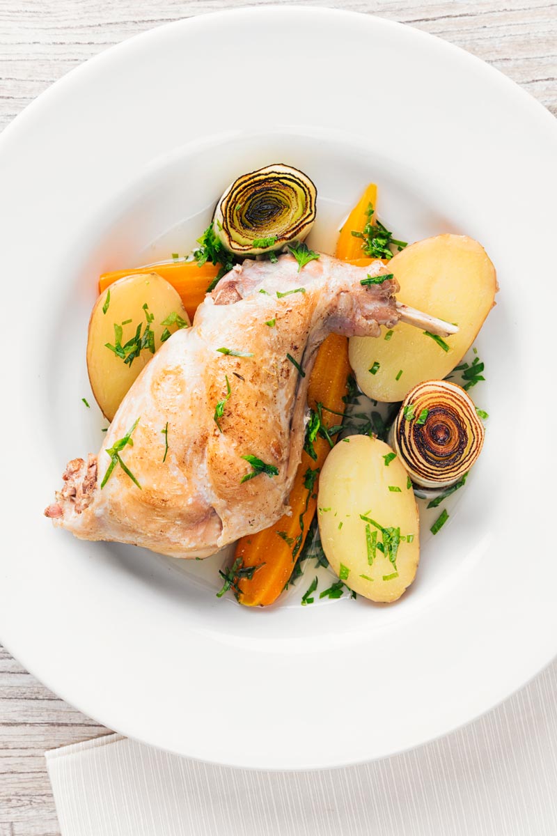Portrait over head image of a braised rabbit leg served with new potatoes, carrots and seared leeks
