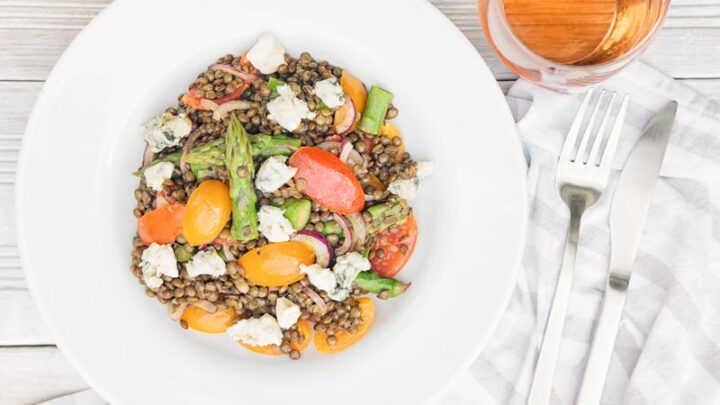 Landscape overhead image of an asparagus salad served in white bowl featuring tomatoes and lentils and crumbled Roquefort cheese