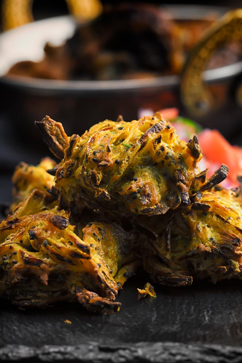 Portrait close up image of onion bhaji pakora served on a slate with a tomato salad and a curry in the background