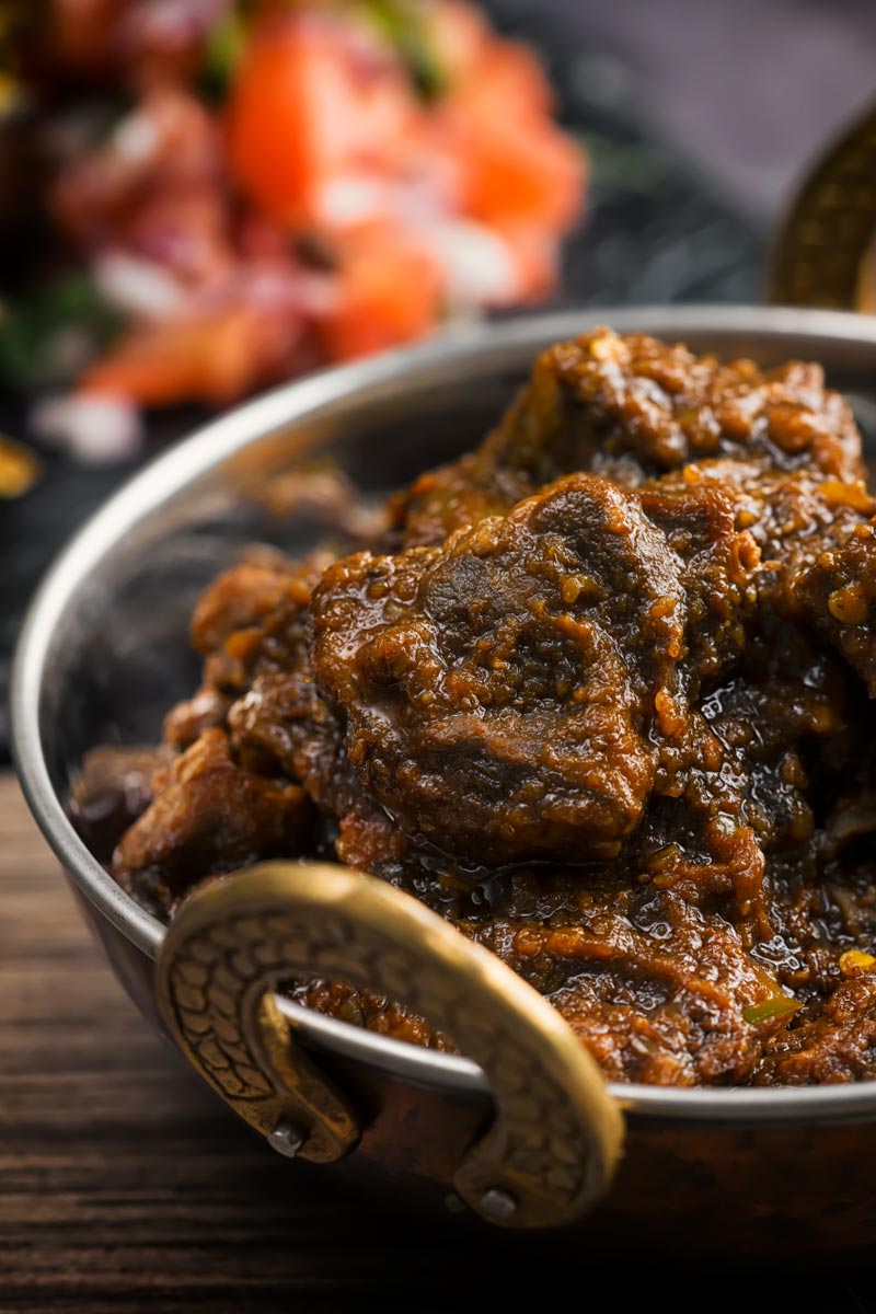 Portrait image of a pork vindaloo curry served in a copper curry bowl