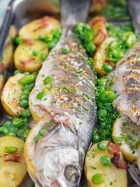 Portrait image of whole baked sea bass in a tray with potatoes peas and broad beans